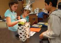 Photo shows three students at a table taking temperature measurements of the tops of foot-high boxes under a heat lamp.