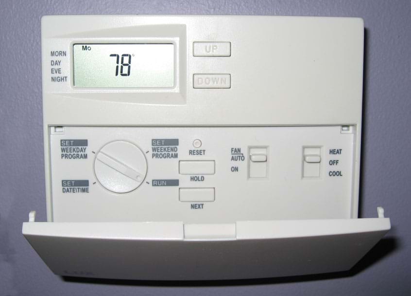 Photo shows an 11 x 9 cm plastic box mounted on a wall, displaying a temperature of 78 &deg;F and many dials and buttons for programming temperature, dates and times.