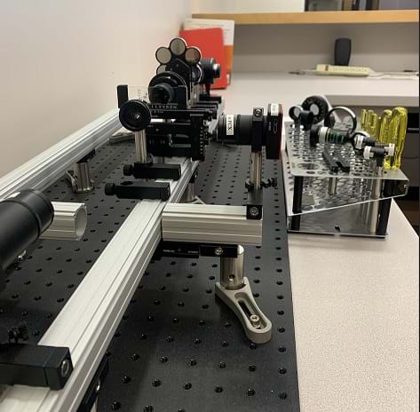 A laser is set up in the bottom left corner facing a series of different optic lenses on a carbon track.  The right side features screwdrivers and tools used to set up the lenses on the track along with additional lenses.