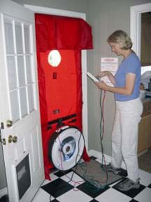 Photo shows a woman standing by a front door where a blower door test is installed for a home energy audit.