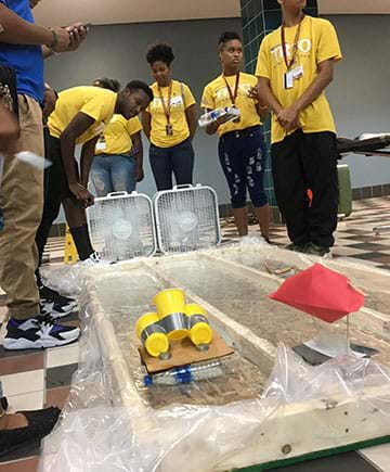 Students stand around two classroom-made water channels and watch their boats race to the opposite end. Two fans propel the boats down each channel.