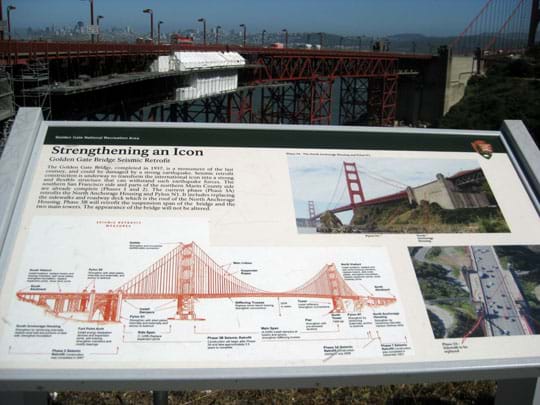 A photograph of a National Park Service sign near the under-construction steel truss and concrete north end of the Golden Gate Bridge in San Francisco. The sign title is "Strengthening an Icon: Golden Gate Bridge Seismic Retrofit," and it shows two photographs and a labeled side-view diagram of the full bridge.