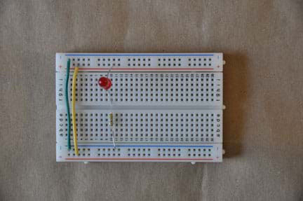 Photo shows a breadboard (circuit board) with the following connections: a yellow wire, which connects the positive terminals on either side of the board; a green wire, which connects the negative terminals on either side of the board; an LED, which is connected to the positive terminal on one side, and the center of the board on the other side; a resistor, which is connected to the same column as the LED in the center of the board, and the negative terminal on the other side.