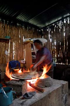 A photograph shows a person cooking tortillas (flatbread) indoors over a wood-fired three-stone stove, essentially an open fire. 
