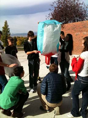 Photo shows 10 students outside, holding a square-shaped green, red and white tissue-paper balloon.