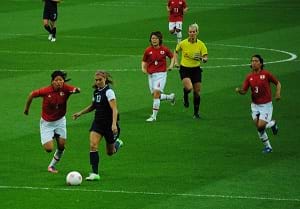A photograph shows a green grassy soccer field with the U.S. women's team playing against the Japanese women's team. Seven players chase a white ball.