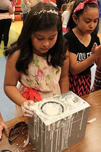 A photograph shows two young girls working with a speaker on a table; the speaker cone is covered with oobleck, a drippy white goo, as are their hands.