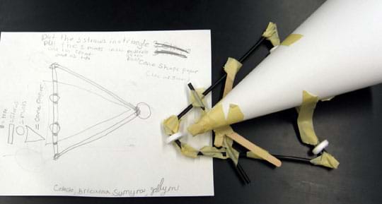 Photo shows a piece of paper with pencil sketches and words next to a vehicle chassis made from black straws, mint candies (wheels), popsicle sticks and tape, with a conical-shaped paper sail.