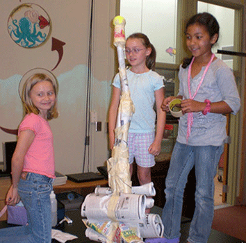 A photograph shows three girls building a tower composed of newspaper, plastic tubs and tape, with a tennis ball on top.  