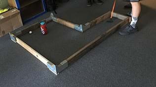 A photograph shows a wooden frame placed on a carpeted floor. The frame edges are made of 2 x 4-inch wood with galvanized metal corner joints as connectors and enclose an L-shaped interior space. A student has just used a golf putter to hit a golf ball from one end of the L shape towards a water bottle (which serves as the hole/goal) in the far side of the L shape. 
