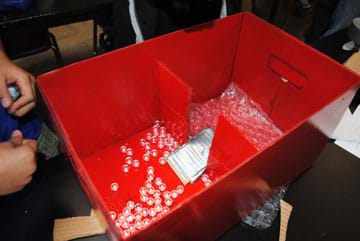 Photo shows a red box with an inner cardboard wall dividing it into two sections. The middle portion of the inner wall contains a one-way valve made in the form of a cardboard and duct tape ramp. Loose marbles are scattered around the floor of both box sections.