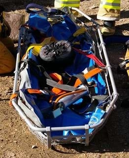 An photograph shows an empty rescue litter up close. Shown is a nonsolid device with a flat surface, similar to a flattened, shallow bowl. Railed sides prevent a person from sliding out of the litter when strapped in (colored security straps shown).