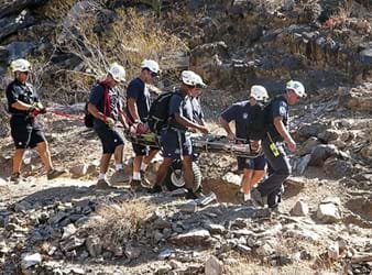 A photograh shows seven people assisting with transporting an injured person down the rocky slope of a mountain. The injured person is on flat surface (similar to a long board) with railed sides; the carrying surface is attached to one large treaded tire, allowing for easier movement over the rough terrain.