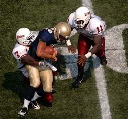 Two football players tackle another football player while wearing padded uniforms on the field during a game.