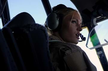A photograph shows a female pilot in the cockpit of an aircraft wearing headphones that include a microphone placed near her mouth as she communicates on the radio.
