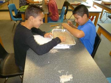 Photo shows two students seated at a table, picking beans out of a bin of sand.