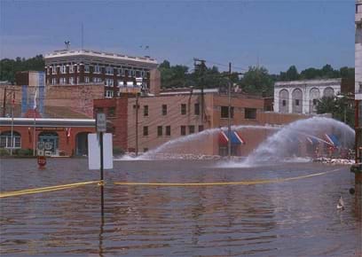 A photograph shows a flooded downtown area with many one to four-story brick buildings. The flood waters reach the bottom of a stop sign. Sprays of water, move water to a different location.