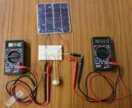 Photo shows two multimeters and a potentiometer on a table hooked up with wires and alligator clips to a circuit board wired to a mini PV panel. Circuit board also wired to round device.
