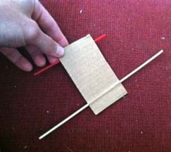 A photograph shows a hand holding a 3 x 5-inch piece of corrugated cardboard with a wooden skewer pushed through the short side via a channel between the cardboard paper layers at one end and a red plastic coffee stirrer inserted through a similar paper channel at the other end. This shows how to place the stirrer axles for the sail car base.
