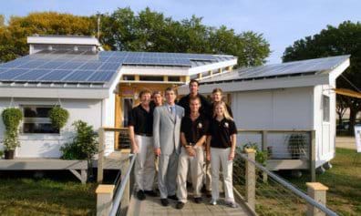 A photo shows a one-story house in which the entire roof surface is angled in one direction and entirely covered with blue solar panels. Five engineering students, their professor and a congressman stand at the entry.