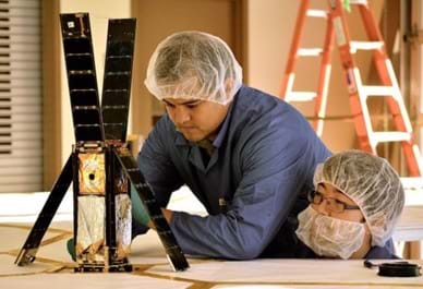 Two men wearing hair and beard nets examine a device on a table. Four solar panel-covered sides of the tall rectangular object (shoe-box sized) are flipped up or away to reveal the inside volume of the satellite cube, which contains a folded-up silver and gold fabric. 