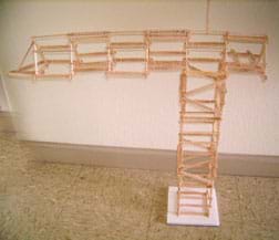 About 100 Popsicle sticks glued end-to-end to create a truss-like tower with a horizontal top structure longer on one side than the other.