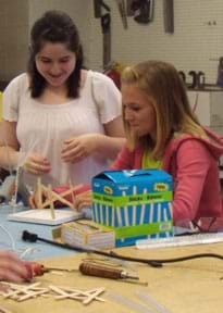 Photo shows two teenage girls gluing Popsicle sticks in a crossed pattern rising from a square foam core board base.
