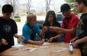 Photo shows five students around a work table, discussing the arrangement for how to glue Popsicle sticks rising up from their square base.