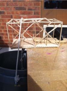 Photo shows a Popsicle stick model truss positioned with half of it extending over the edge of a table. The truss is secured to the table using a clamp and a piece of wood. A bucket is hung from the cantilevered end by a rope.