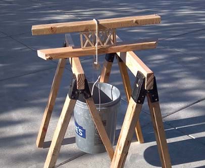 Photo shows a truss sandwiched between two 2x4s, and the sandwich is placed across a set of saw horses. The bucket is hung below the truss by looping the rope over the sandwiched truss.