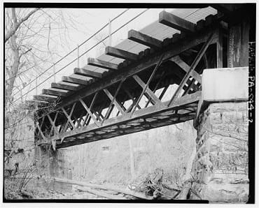 A black and white image of an old railroad bridge with trusses under the deck.