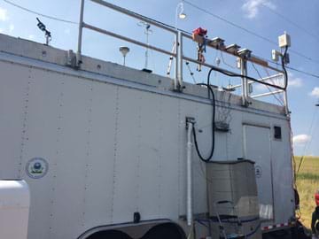 A photograph shows a white box-shaped trailer on wheels with railings around the rooftop onto which equipment is attached, including monitors, sensors, hoses and inlets into the trailer. 