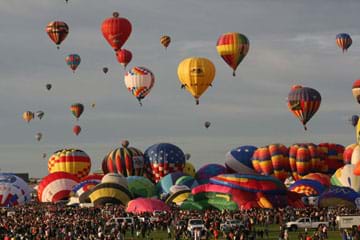 Photo shows a park filled with people and about 20 colorful hot air balloons on the ground. Another 22 hot air balloons are floating in the sky.