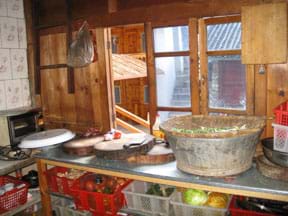 A waist-high kitchen counter against a wall holds cutting boards, knife and containers. Shelves below hold bins of fruits and vegetables. The wall is made of wooden boards with an operable two-part window.