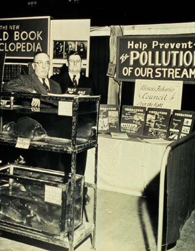 A black/white photograph of two men standing in an exhibit booth with signs such as, "Help Prevent the Pollution of Our Streams."