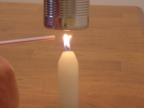 A photograph shows a candle burning below a soup can while someone uses a straw to gently blow the flame away from the bottom of the can, preventing the buildup of carbon.