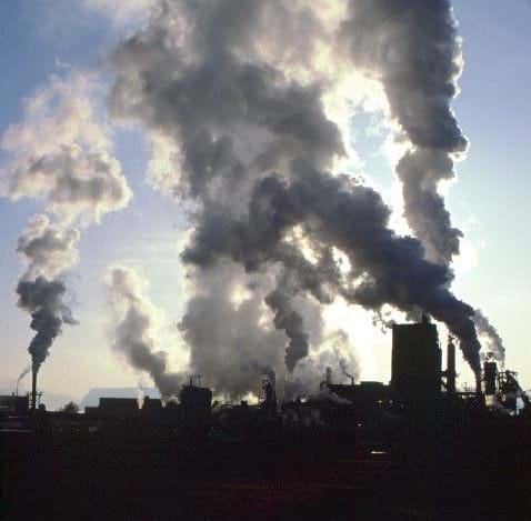 A photograph of industrial factories releasing pollutants. A grayish haze around the factory indicates the presence of industrial smog.