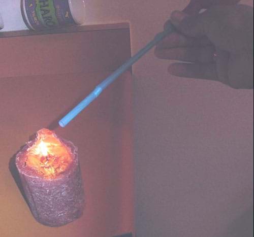 A photograph shows a tilted bottle with a plastic drinking straw attached to its mouth. The other end of the straw is positioned near the flame of a burning candle.