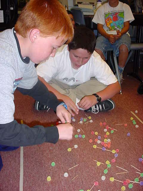 Two students sitting on the floor surrounded by a scattering of toothpicks and gumdrops.