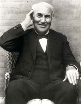 Black and white photo shows a seated and smiling Thomas Edison cupping his right hand around his right ear.