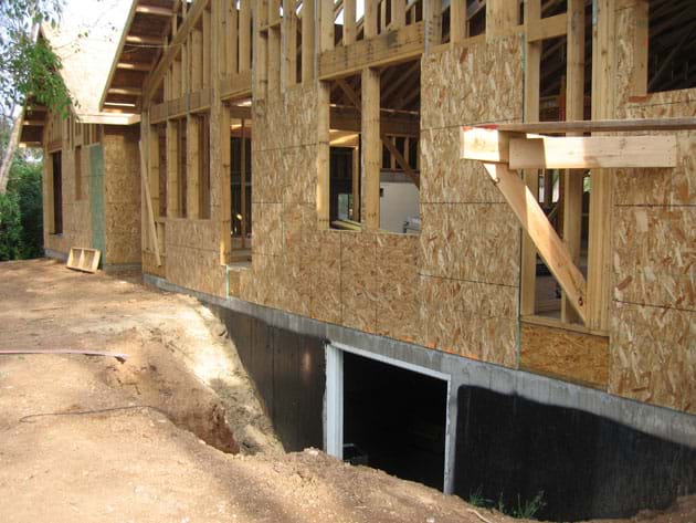 Photo shows an unfinished wood-framed one-story house, with some of its concrete foundation (basement walls) exposed (not backfilled).