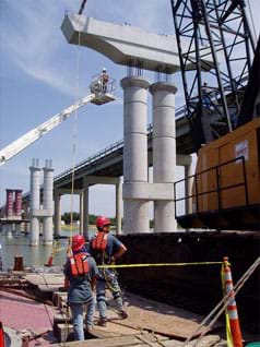 Construction site photo shows a crane placing a beam of concrete atop two concrete columns.