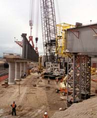 Photo shows a construction site with steel and concrete members, cranes, equipment and workers in hard hats.