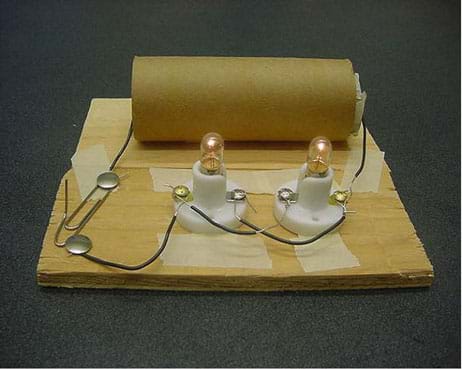 A photograph of a parallel circuit made with two D-cell batteries, two light bulb holders, two light bulbs and a switch. The two D-cell batteries are contained within a cardboard paper towel tube. One wire leads from the tube to the switch, which is made from two thumbtacks and a paper clip. Another wire exits the opposite end of the tube and is connected to one of the light bulb holders. The two light bulb holders are connected in parallel using short wires. The switch is closed and the two light bulbs are lit.
