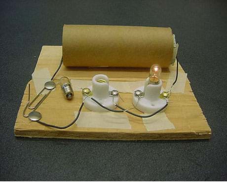A photograph of a parallel circuit made with two D-cell batteries, two light bulb holders, two light bulbs and a switch. The two D-cell batteries are contained within a cardboard paper towel tube. One wire leads from the tube to the switch, which is made from two thumbtacks and a paper clip. Another wire exits the opposite end of the tube and is connected to one of the light bulb holders. The two light bulb holders are connected in parallel using short wires. The switch is closed. Although one light bulb is removed from its holder, the remaining light bulb is still lit.