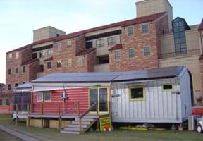 Photograph of a modern mobile-home-size structure in front of a huge university building.