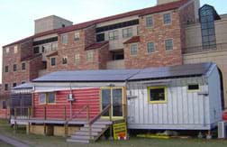 A photograph shows a modern mobile-home-size structure in front of a huge university building.
