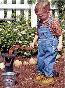 Photo shows a younge boy in denim overalls and work boots, pumping water from an old-fashioned well.