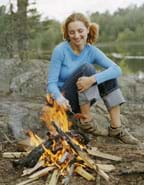 Photo shows a girl roasting a hot dog in the flames of a campfire of burning wood.