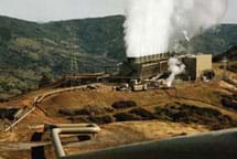 Photo shows a large building on a hilltop with many pipes to it and water vapor emissions leaving the structure.